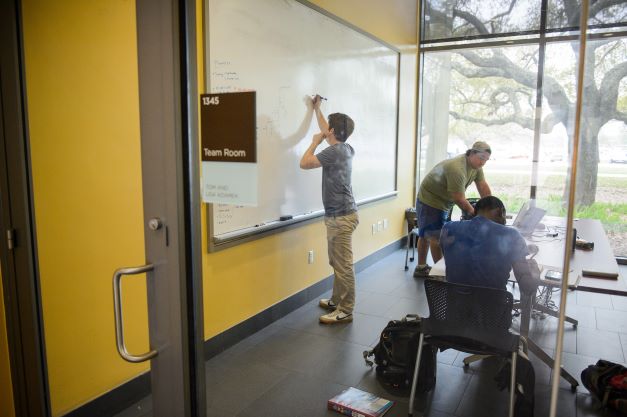student writes on white board while two other students make notes at a desk