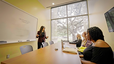 Monique Domingue, a faculty member, stands at a whiteboard and speaks to a small group of students. She has a warm, excited expression.