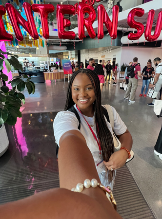 Photo of a female student taking a selfie at her internship site, Adobe. There are red letter balloons in the background that read Summer Interns.