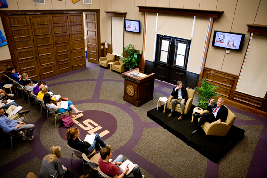 Picture of a forum space with students in the audience and two men sitting in arm chairs in the front of room speaking to one another
